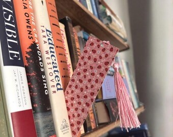 Può includere: Un segnalibro rosa con un motivo a coccinella e un fiocco rosa. Il segnalibro è su una libreria con diversi libri.