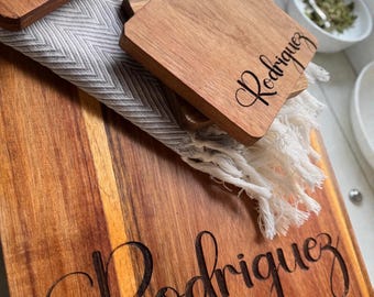 Può includere: Tagliere e sottobicchieri in legno con il nome "Rodriguez" inciso in corsivo. Il tagliere è rettangolare con venature naturali del legno. I sottobicchieri sono quadrati e impilati su un panno grigio e bianco.