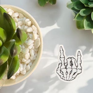 May include: A white sticker featuring a black outline of a skeleton hand in a rock and roll pose. The sticker is on a white surface, next to a small succulent plant in a beige pot filled with white pebbles.