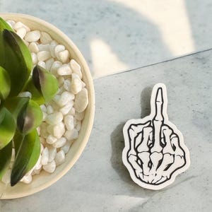 May include: A white sticker with a black outline of a skeleton hand giving the middle finger. The sticker is on a light gray surface, next to a small beige bowl filled with white pebbles and a green plant.