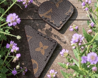 Può includere: Due sottobicchieri in pelle marrone, uno a forma di cuore e uno triangolare, con un motivo dorato ripetuto. I sottobicchieri sono su una superficie di legno invecchiato, circondati da fiori viola e fogliame verde.