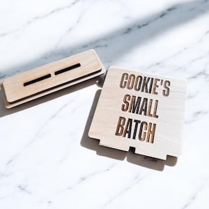 May include: Two wooden cookie box pieces. One rectangular piece with two slots and another square piece with the words "COOKIE'S SMALL BATCH" etched into the wood. The pieces are light brown and sit on a white marble surface.