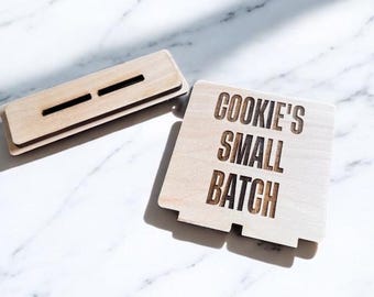 Può includere: Due pezzi di legno per scatola di biscotti. Un pezzo rettangolare con due fessure e un altro pezzo quadrato con le parole "COOKIE'S SMALL BATCH" incise nel legno. I pezzi sono marrone chiaro e poggiano su una superficie di marmo bianco.