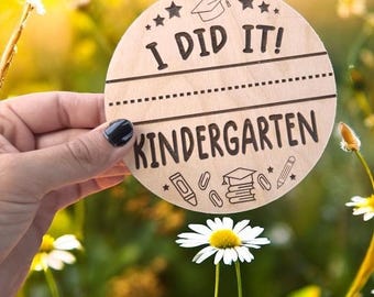 Può includere: Un disco di legno con le parole "I DID IT! KINDERGARTEN" in nero. Il disco presenta un cappello di laurea, stelle e illustrazioni di articoli scolastici. Lo sfondo è un campo di margherite.