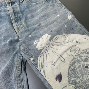 May include: Light blue denim jeans with a floral and decorative print. The jeans feature a button fly and pockets. The print includes white flowers, purple leaves, and other decorative elements. The jeans have a faded wash.