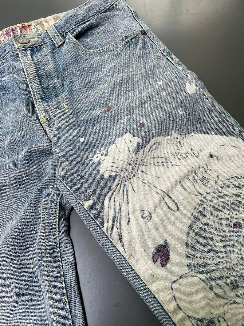 May include: Light blue denim jeans with a floral and decorative print. The jeans feature a button fly and pockets. The print includes white flowers, purple leaves, and other decorative elements. The jeans have a faded wash.
