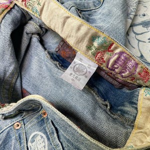 May include: Light blue denim jeans with decorative embroidered waistband and pocket details. The waistband features floral and geometric patterns in red, purple, and green. The jeans have a white embroidered logo on the pocket.