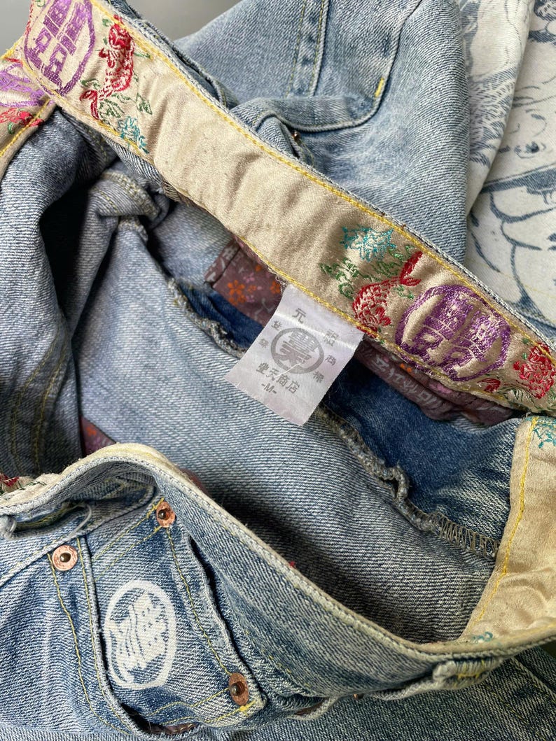 May include: Light blue denim jeans with decorative embroidered waistband and pocket details. The waistband features floral and geometric patterns in red, purple, and green. The jeans have a white embroidered logo on the pocket.