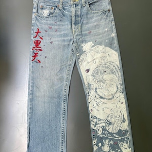 May include: Light blue denim jeans with a faded wash. The jeans feature a large white graphic print on the right leg and red Japanese text on the left leg. Small purple accents are scattered across the front.