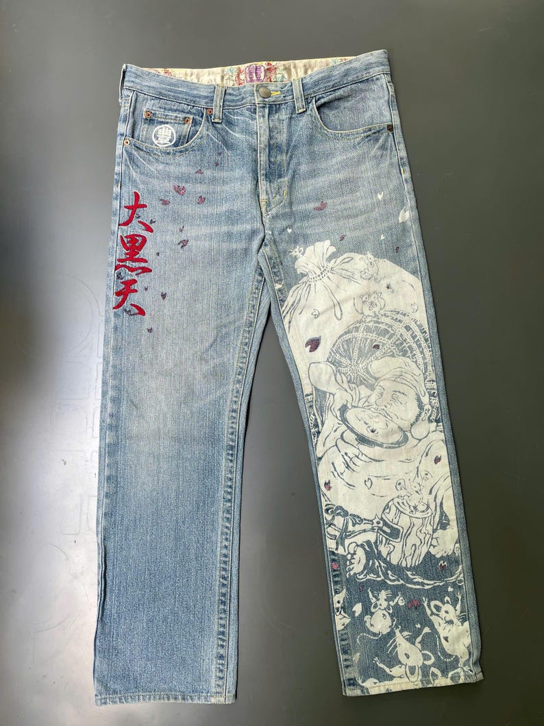 May include: Light blue denim jeans with a faded wash. The jeans feature a large white graphic print on the right leg and red Japanese text on the left leg. Small purple accents are scattered across the front.
