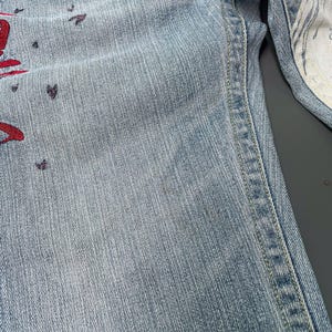 May include: Light blue denim jeans with red and purple decorative accents. The jeans have a classic five-pocket design and a straight leg cut. The interior lining of the jeans is white with a patterned design.