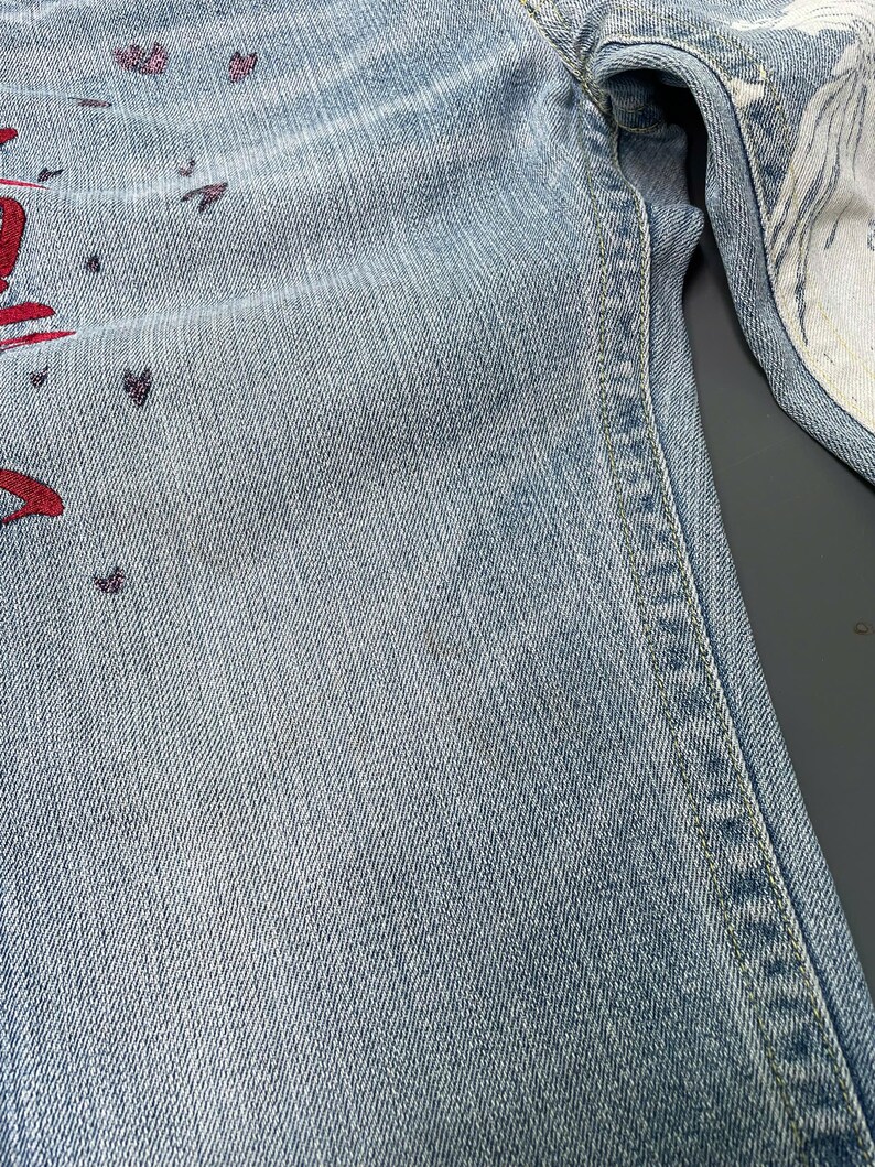 May include: Light blue denim jeans with red and purple decorative accents. The jeans have a classic five-pocket design and a straight leg cut. The interior lining of the jeans is white with a patterned design.