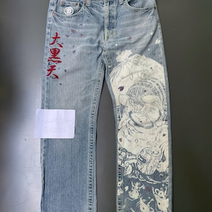 May include: Light blue denim jeans with a unique design. The jeans feature a large white and blue illustration on the right leg and red Japanese characters on the left leg. The jeans have a button fly and a patterned waistband.