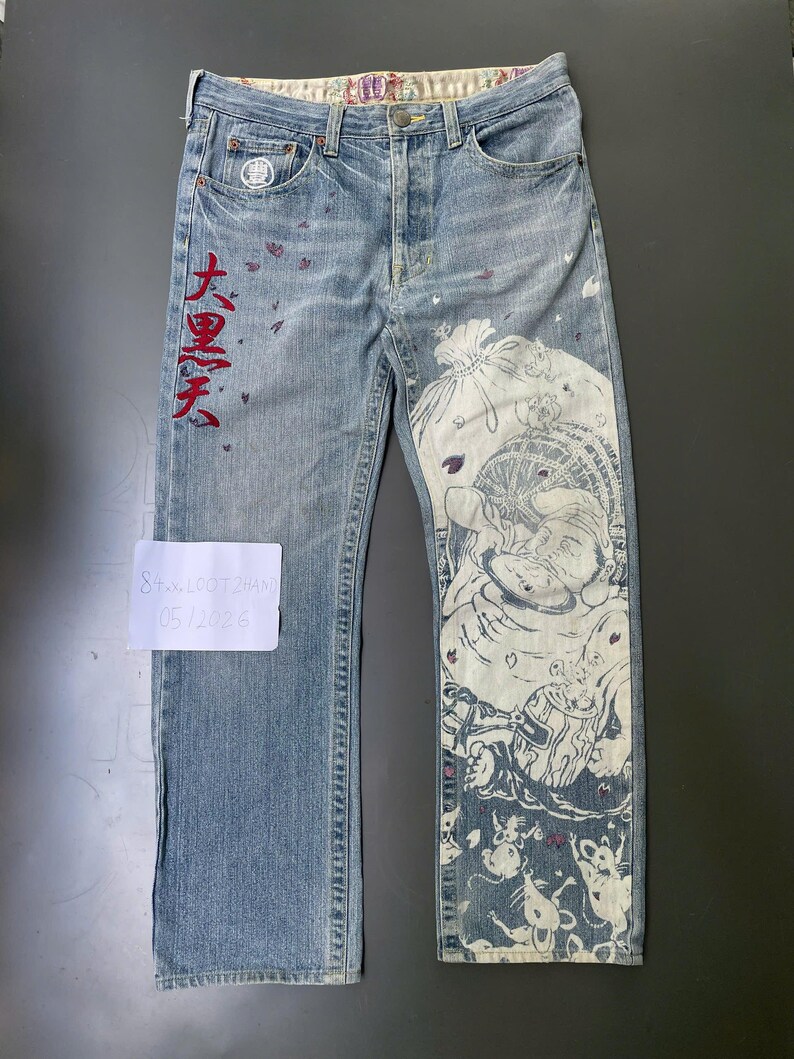 May include: Light blue denim jeans with a unique design. The jeans feature a large white and blue illustration on the right leg and red Japanese characters on the left leg. The jeans have a button fly and a patterned waistband.