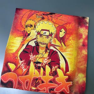 May include: A tie-dye t-shirt featuring a vibrant orange and yellow graphic print of Naruto Uzumaki, Minato Namikaze, and Kushina Uzumaki. The design includes a large red fox silhouette in the background and Japanese text at the bottom of the shirt.