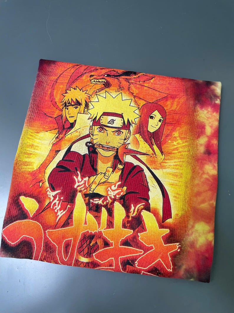 May include: A tie-dye t-shirt featuring a vibrant orange and yellow graphic print of Naruto Uzumaki, Minato Namikaze, and Kushina Uzumaki. The design includes a large red fox silhouette in the background and Japanese text at the bottom of the shirt.