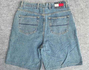 May include: Light blue denim shorts featuring a high-waisted design with belt loops, two back pockets with horizontal stitching, and a rectangular Tommy Jeans brand patch in red, white, and blue located on the back right side of the waistband.