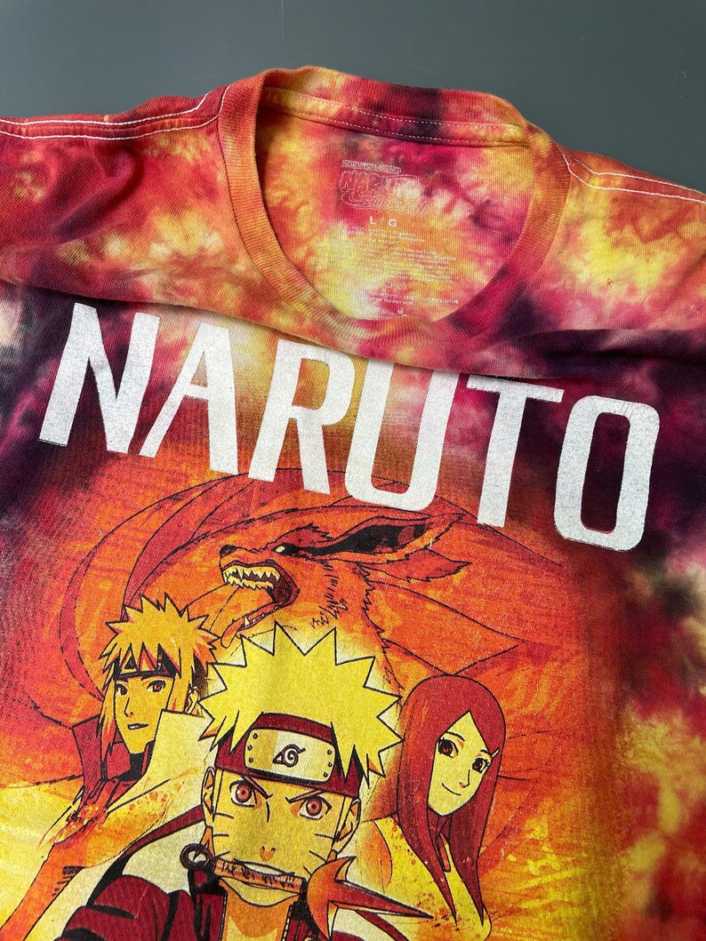 May include: A tie-dye t-shirt in shades of red, orange, and yellow featuring the word NARUTO in large white letters. Below the text is a graphic illustration of three anime characters and a large fox creature with multiple tails in the background.