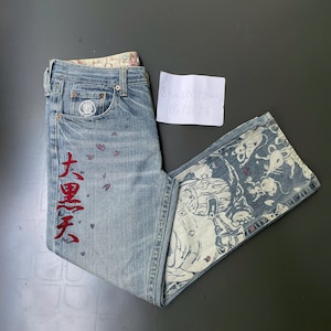 May include: Light blue denim jeans with red Japanese characters and small red butterfly accents. The jeans feature a decorative patterned inner lining and a unique design on the flared leg. A small white tag is visible.
