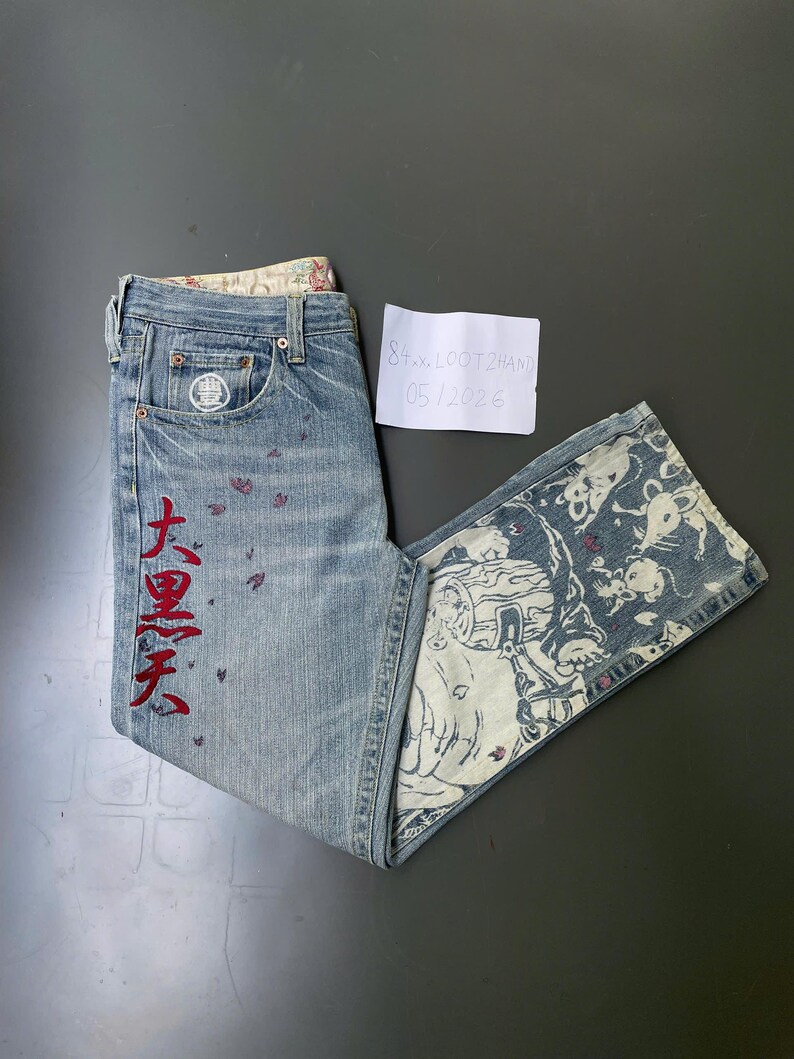 May include: Light blue denim jeans with red Japanese characters and small red butterfly accents. The jeans feature a decorative patterned inner lining and a unique design on the flared leg. A small white tag is visible.