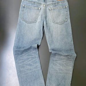 May include: Light blue denim jeans with a classic five-pocket design. The jeans have a straight leg cut and a faded wash, with a leather patch on the back waistband. The jeans are laid flat on a dark surface.