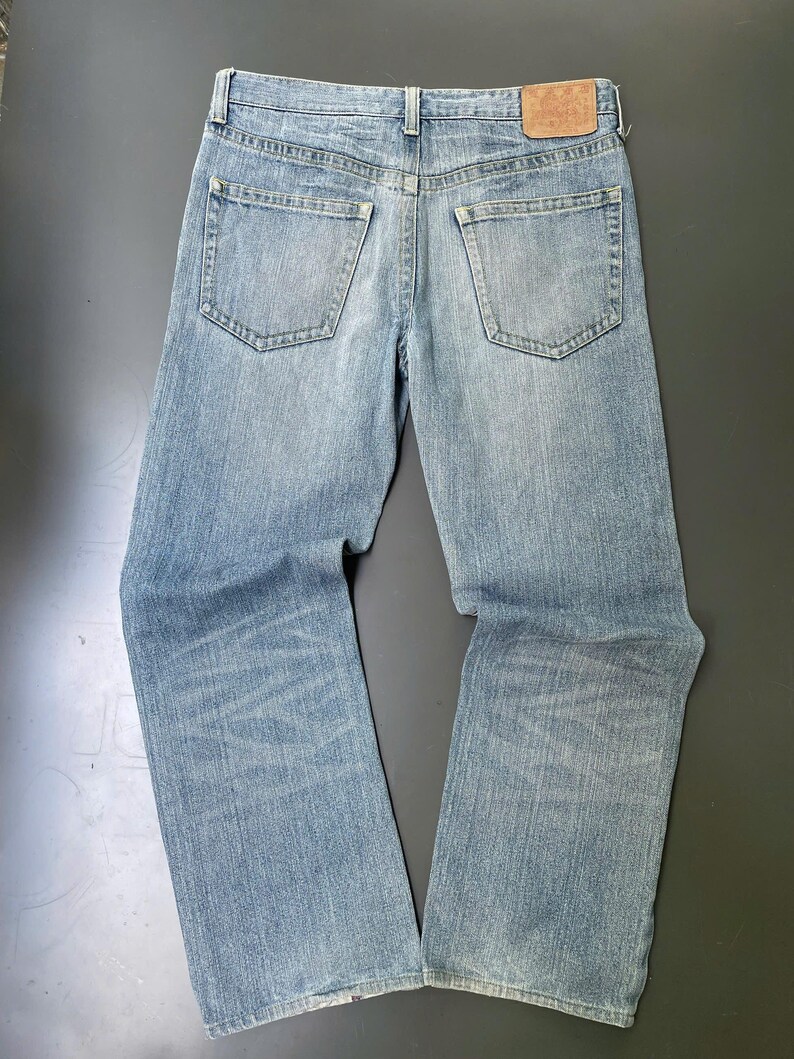 May include: Light blue denim jeans with a classic five-pocket design. The jeans have a straight leg cut and a faded wash, with a leather patch on the back waistband. The jeans are laid flat on a dark surface.