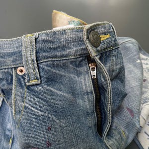 May include: Close-up of blue denim jeans with a button closure and a zipper. The jeans have decorative stitching in yellow and red accents. The waistband features a patterned fabric lining.