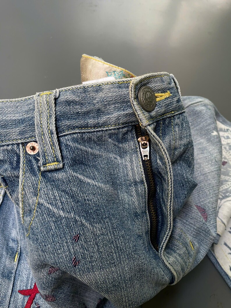 May include: Close-up of blue denim jeans with a button closure and a zipper. The jeans have decorative stitching in yellow and red accents. The waistband features a patterned fabric lining.