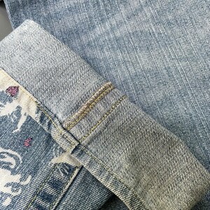 May include: Close-up of light blue denim jeans with a folded cuff. The cuff reveals a patterned fabric with white and pink floral designs. The jeans have visible stitching and a faded appearance.