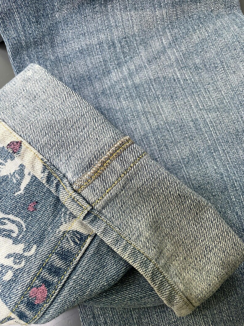 May include: Close-up of light blue denim jeans with a folded cuff. The cuff reveals a patterned fabric with white and pink floral designs. The jeans have visible stitching and a faded appearance.