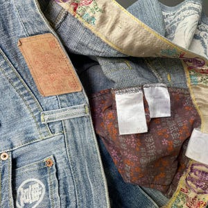 May include: Close-up of light blue denim jeans with decorative floral embroidery on the inner lining and waistband. Features a brown leather patch and a white logo on the pocket. The jeans have metal rivets and a button closure.