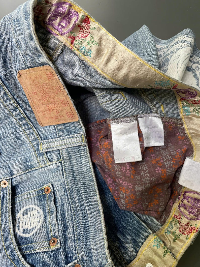 May include: Close-up of light blue denim jeans with decorative floral embroidery on the inner lining and waistband. Features a brown leather patch and a white logo on the pocket. The jeans have metal rivets and a button closure.