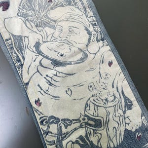 May include: Close-up of denim jeans with a detailed illustration. The design features a figure with a hat, holding a plate, and a beer stein. The artwork is primarily white and blue, with accents of pink. The jeans have a classic five-pocket design.