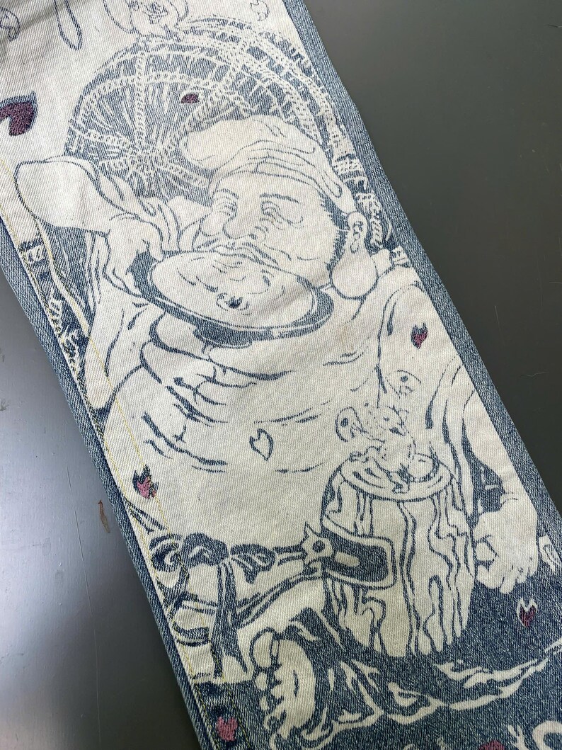 May include: Close-up of denim jeans with a detailed illustration. The design features a figure with a hat, holding a plate, and a beer stein. The artwork is primarily white and blue, with accents of pink. The jeans have a classic five-pocket design.