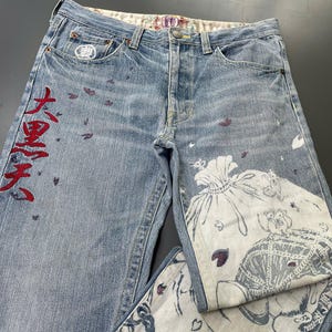 May include: Light blue denim jeans with a button fly and decorative stitching. The jeans feature red Japanese characters and a white and blue graphic print on the front and back pockets. Scattered purple accents add detail.
