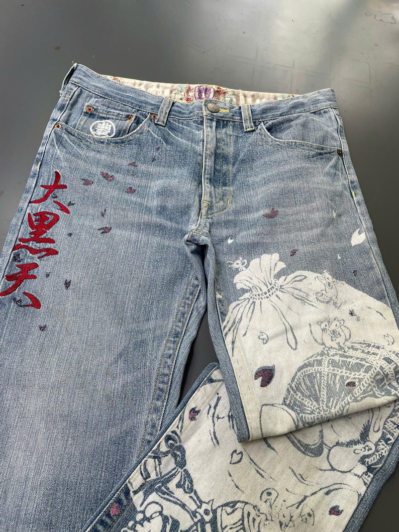 May include: Light blue denim jeans with a button fly and decorative stitching. The jeans feature red Japanese characters and a white and blue graphic print on the front and back pockets. Scattered purple accents add detail.