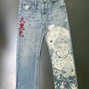 May include: Light blue denim jeans with a faded wash. The jeans feature a large white graphic print on the right leg and red Japanese text on the left leg. Small purple accents are scattered across the front.