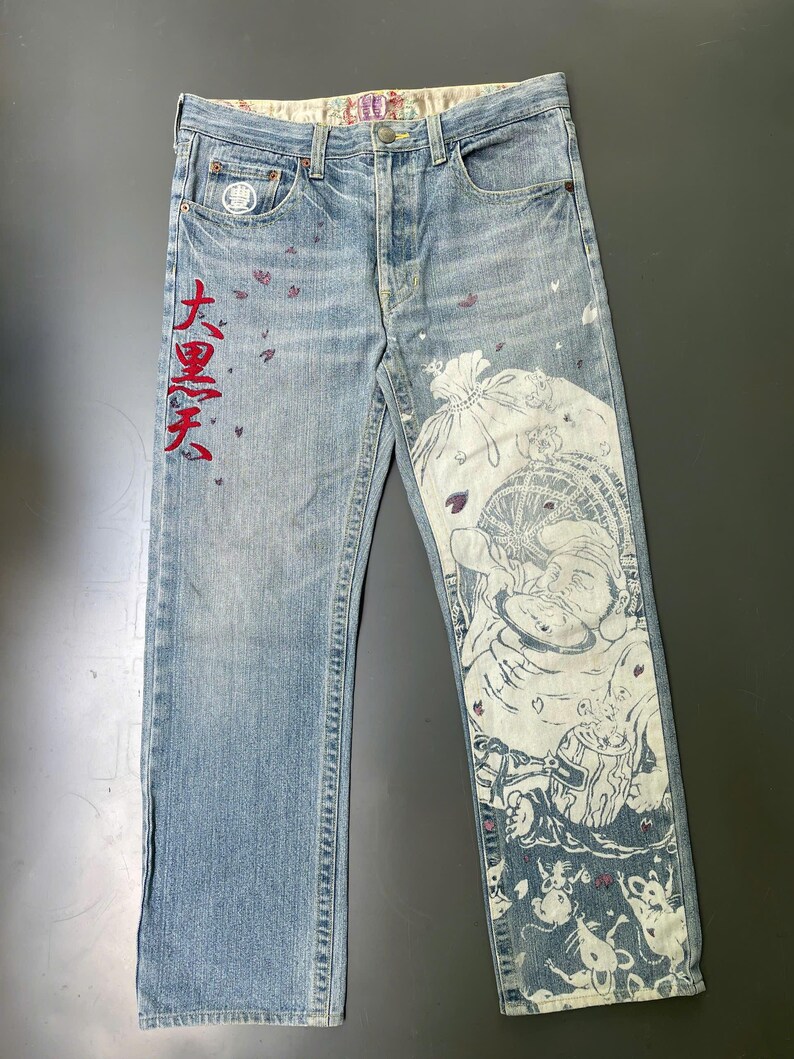 May include: Light blue denim jeans with a faded wash. The jeans feature a large white graphic print on the right leg and red Japanese text on the left leg. Small purple accents are scattered across the front.