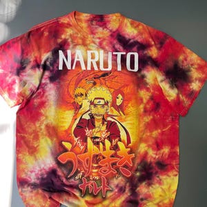 May include: A short-sleeve t-shirt with a red, orange, and yellow tie-dye pattern. The front features the text NARUTO in white, a graphic of Naruto Uzumaki with other characters, and Japanese text in red and yellow at the bottom of the design.