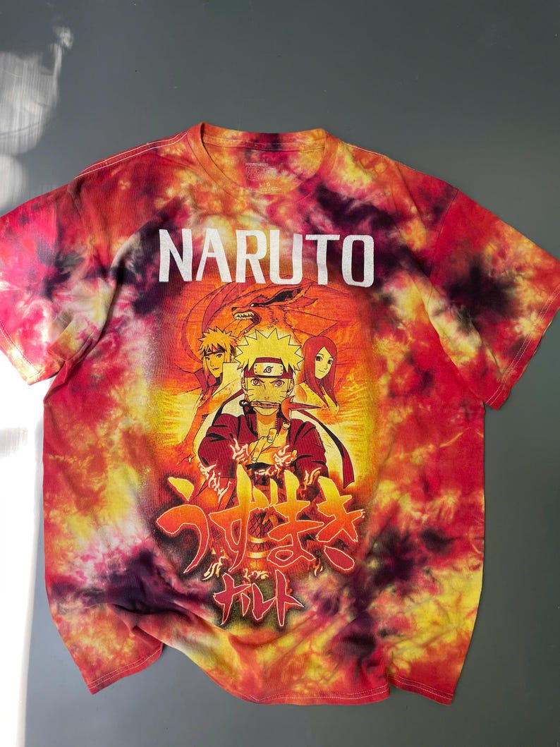 May include: A short-sleeve t-shirt with a red, orange, and yellow tie-dye pattern. The front features the text NARUTO in white, a graphic of Naruto Uzumaki with other characters, and Japanese text in red and yellow at the bottom of the design.