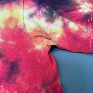 May include: A short-sleeved t-shirt featuring a vibrant tie-dye pattern in shades of bright pink, deep purple, and pale yellow. The garment displays a visible seam along the sleeve and underarm area, showcasing the blended color transitions.