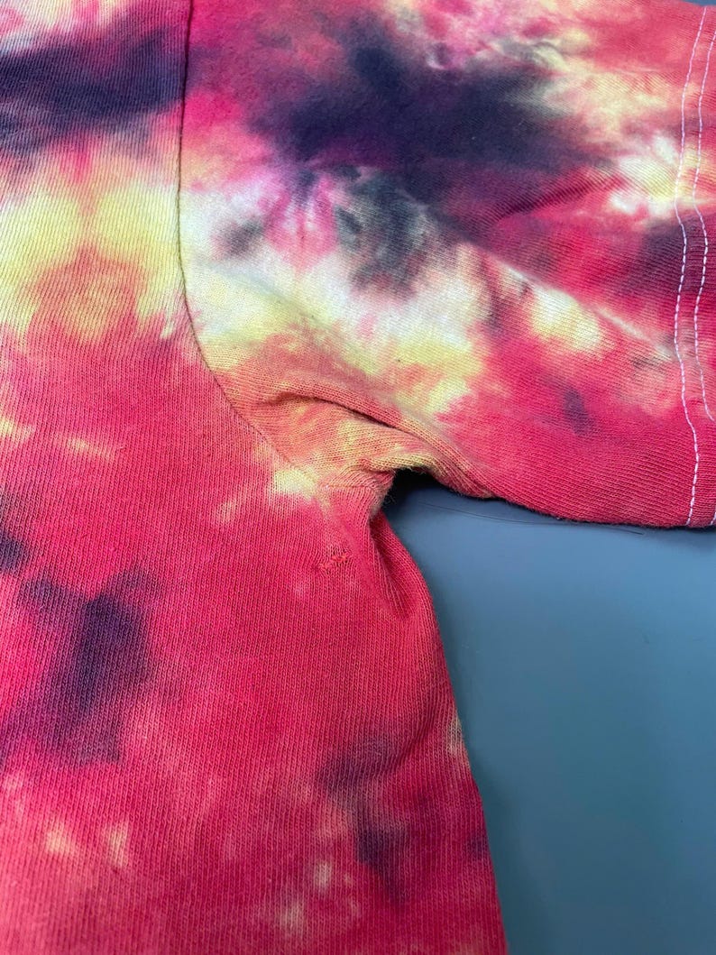 May include: A short-sleeved t-shirt featuring a vibrant tie-dye pattern in shades of bright pink, deep purple, and pale yellow. The garment displays a visible seam along the sleeve and underarm area, showcasing the blended color transitions.