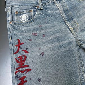 May include: Light blue denim jeans with red Japanese characters embroidered down the side. The jeans feature a white logo on the front pocket and small purple floral accents. The interior waistband has a floral pattern.