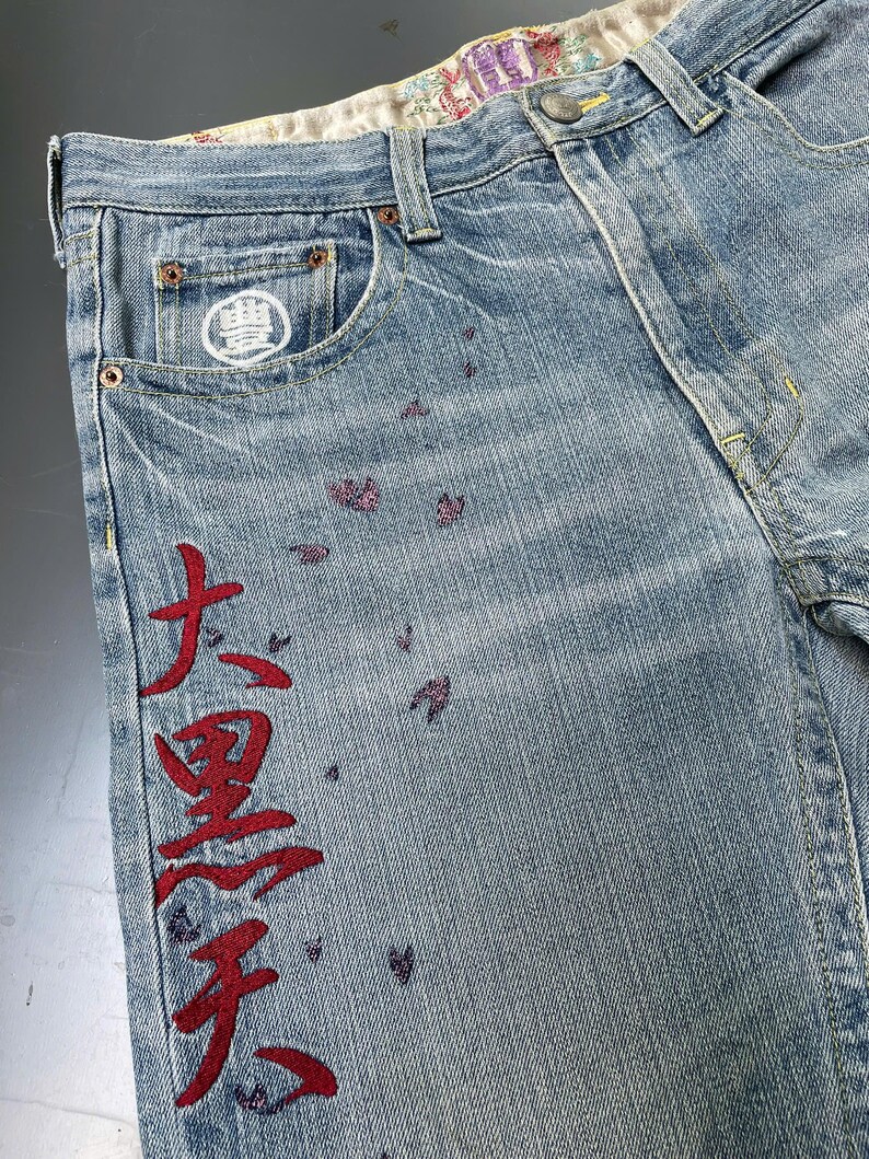 May include: Light blue denim jeans with red Japanese characters embroidered down the side. The jeans feature a white logo on the front pocket and small purple floral accents. The interior waistband has a floral pattern.