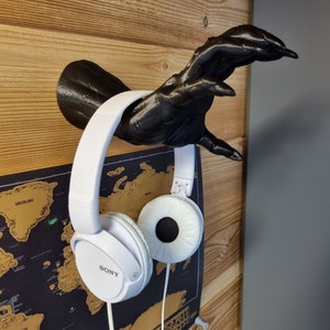 Peut inclure: Casque audio blanc avec une main noire imprimée en 3D qui le tient en l'air. Le casque porte l'inscription "SONY" sur le côté. Le casque est accroché à un mur en bois avec une carte du monde derrière.