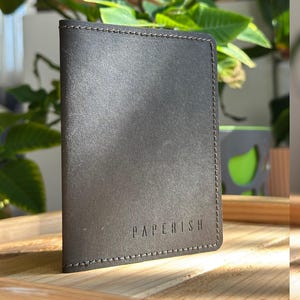 Puede incluir: Una cartera de cuero gris oscuro con la palabra "PAPERISH" grabada en la parte delantera. La cartera tiene un borde cosido y se muestra sobre una superficie de madera. El fondo incluye plantas verdes y un vistazo a un objeto decorativo.
