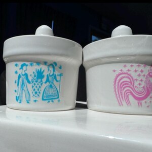 May include: Two white ceramic canisters with lids. The left canister features a blue graphic of two figures and a plant. The right canister features a pink graphic of a stylized rooster surrounded by small star shapes. Both have a smooth finish.