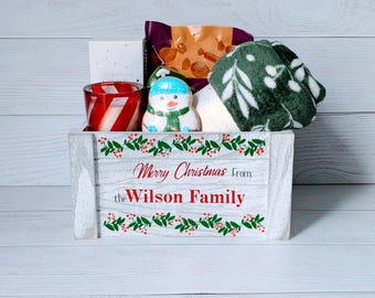 May include: A white wooden crate filled with Christmas gifts. The crate is decorated with green leaves and red berries, and the text "Merry Christmas From the Wilson Family" is printed on the front. Inside are a snowman, a candle, and a pair of green mittens.