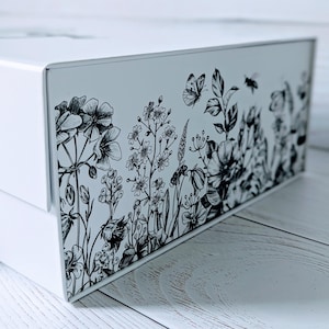 May include: A white rectangular box with a black and white floral design. The design features detailed illustrations of flowers, butterflies, and bees. The box is set against a light-coloured wooden surface.