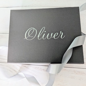 May include: A black gift box with a silver ribbon tied around it. The box has the name "Oliver" printed on the front in white.